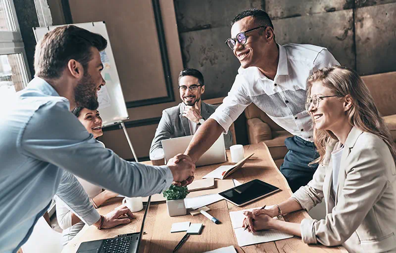 Two business people shake hands while their team smiles.